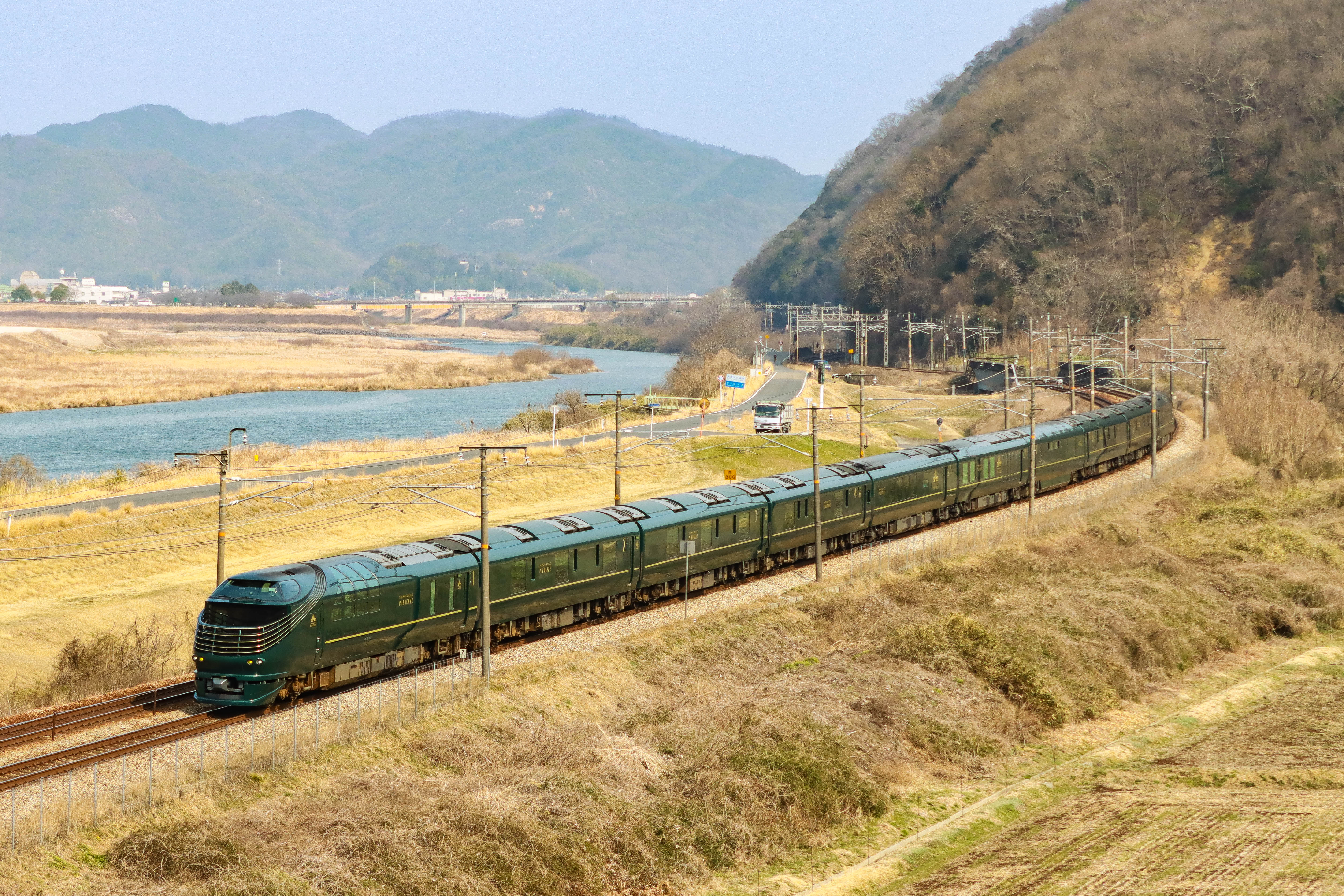 The green TWILIGHT EXPRESS MIZUKAZE cruise train running beside a river valley on the San’yo main line.
