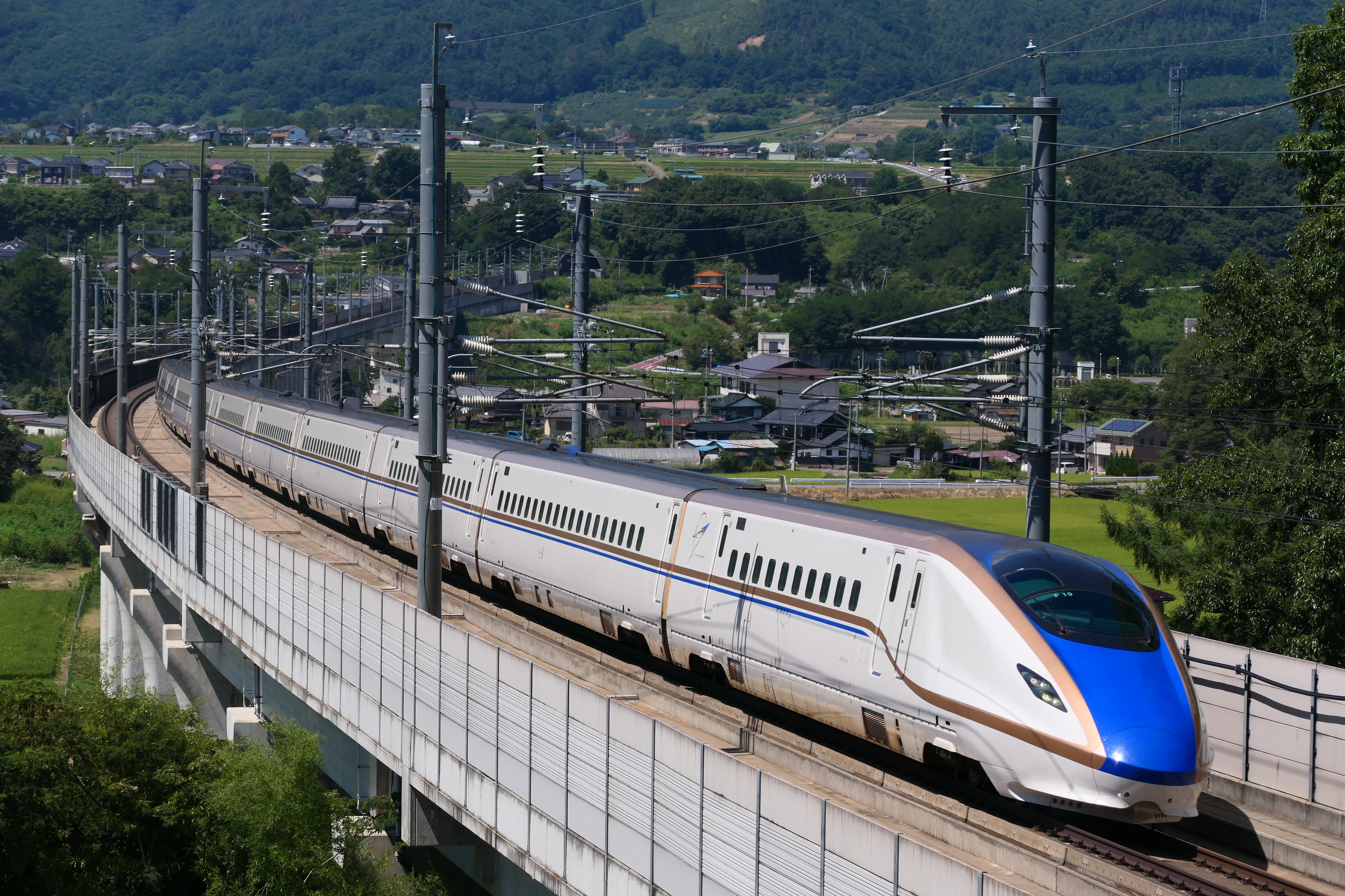 An E7 series Hokuriku Shinkansen train crossing a viaduct above a rural valley.