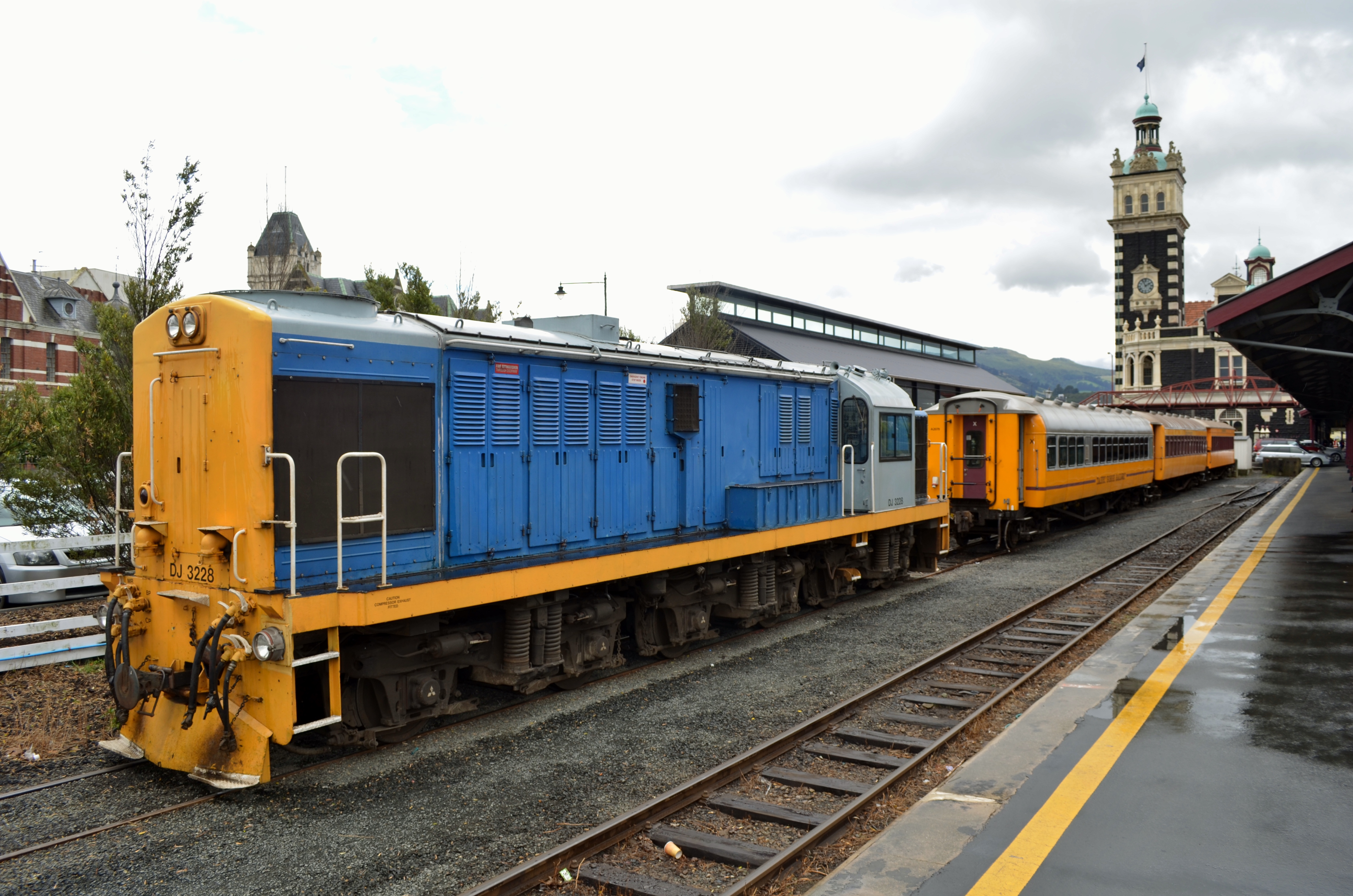 Locomotive DJ3228 alongside a Taieri Gorge Railway train at Dunedin Railway Station, the station clock tower behind.