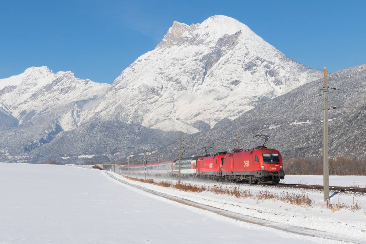 A passenger train crossing the Arlberg Railway near Flaurling in Tyrol, Austria, with Hohe Munde in the background.