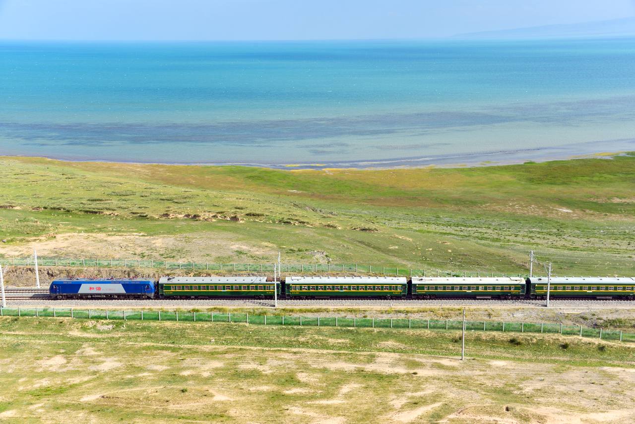 A green and yellow Chinese passenger train running beside Qinghai Lake on the Qinghai-Tibet Railway.